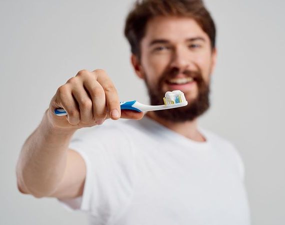 Junger Mann mit Bart hält vor sich die Zahnbürste mit Zahnpasta drauf und lächelt.