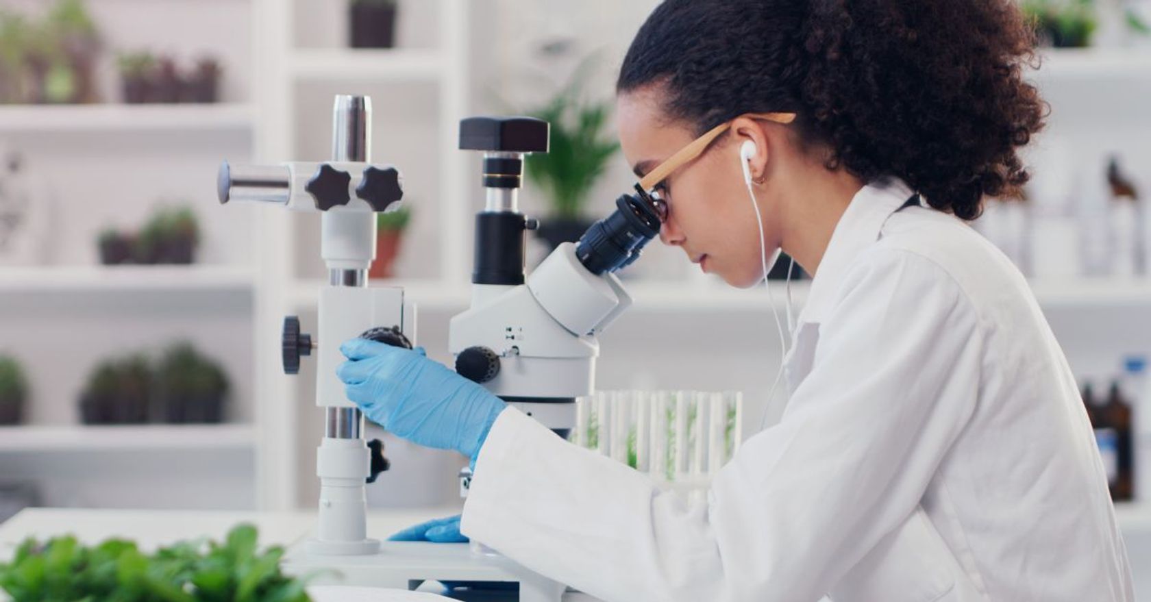 Junge Frau im Labor blickt durch ein Mikroskop, im Vordergrund und Hintergrund sind Pflanzen zu sehen.