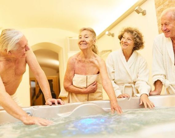 Eine Gruppe Senioren wartet fröhlich auf ihre Hydrotherapie. Sie halten schonmal die Hand ins Wasser der Badewanne und lachen.