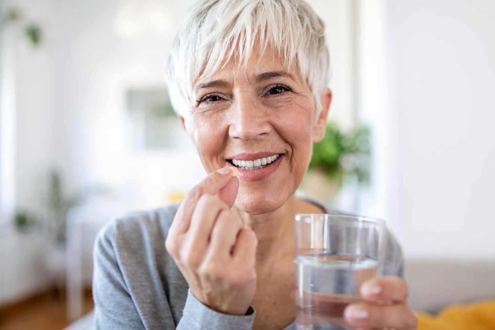 Hübsche Seniorin mit Kurzhaarschnitt hält eine Vitaminpille und ein Glas Wasser in den Händen und lächelt.