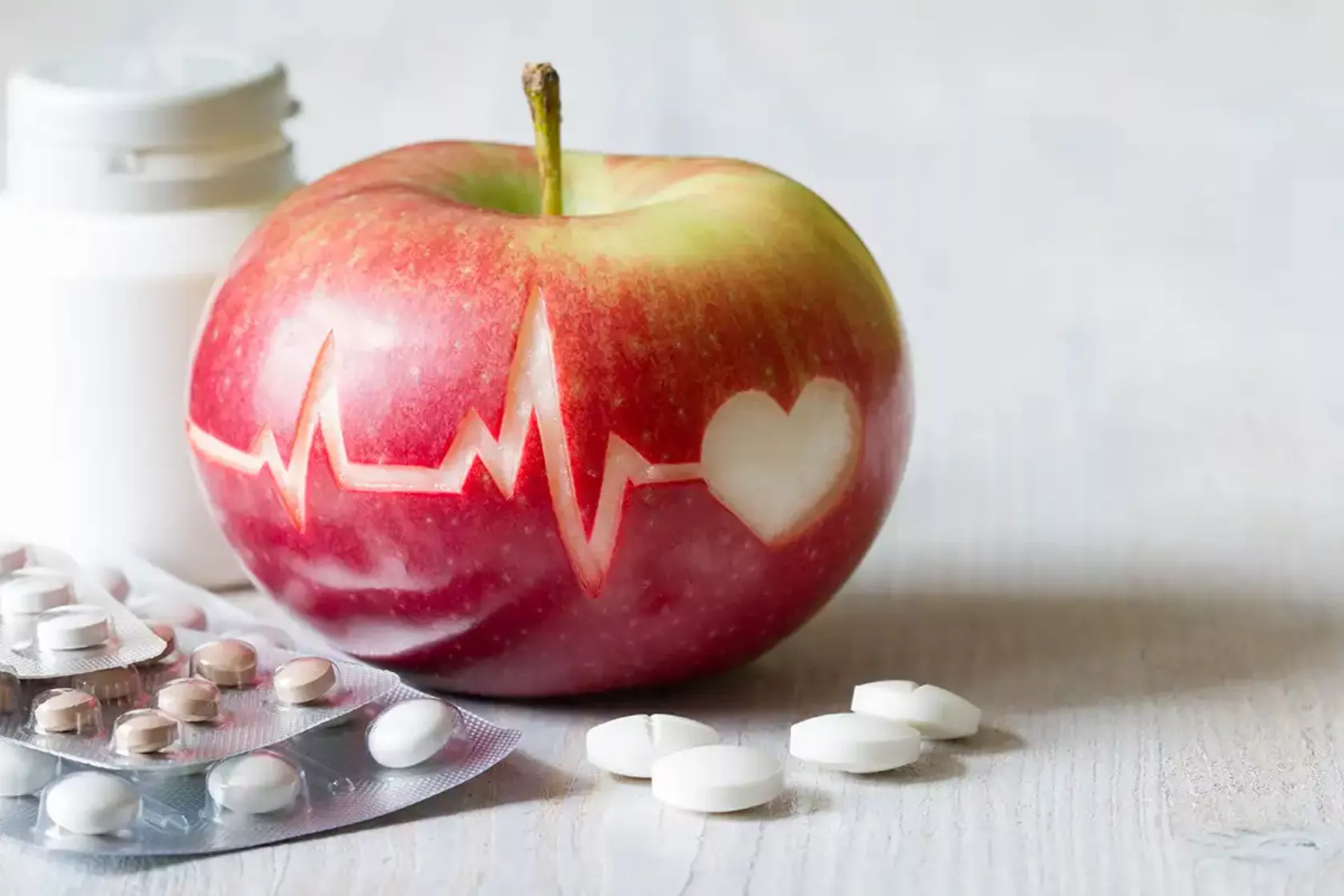 In einen Apfel ist eine EKG-Kurve eingeritzt mit einem Herz, drumherum liegen Tabletten, Dragees, Kapseln.
